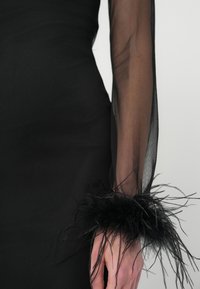 Vestido preto justo com mangas longas transparentes, apresentando um dramático detalhe em penas pretas nos punhos. Textura suave e silhueta slim.