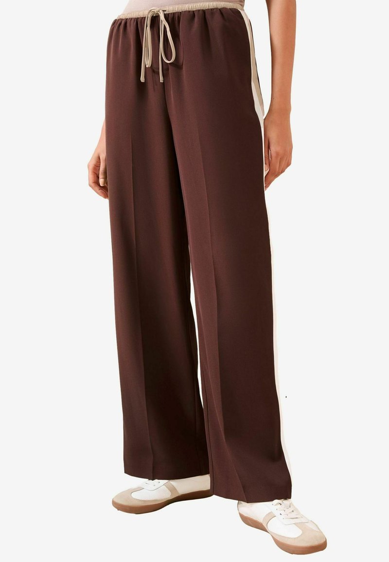Bruine wijde broek met beige trekkoord in de taille en zijkanten in een andere kleur, gedragen met wit en beige sneakers.