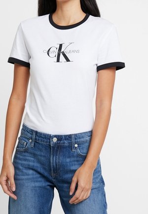 Biała koszulka z krótkim rękawem z czarną lamówką i logo „Calvin Klein Jeans” zestawiona z niebieskimi dżinsami na stojącej osobie.