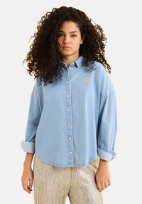 Camicia in denim azzurro chiaro con bottone, dal taglio morbido, maniche arrotolate e un piccolo dettaglio ricamato in oro sul petto.