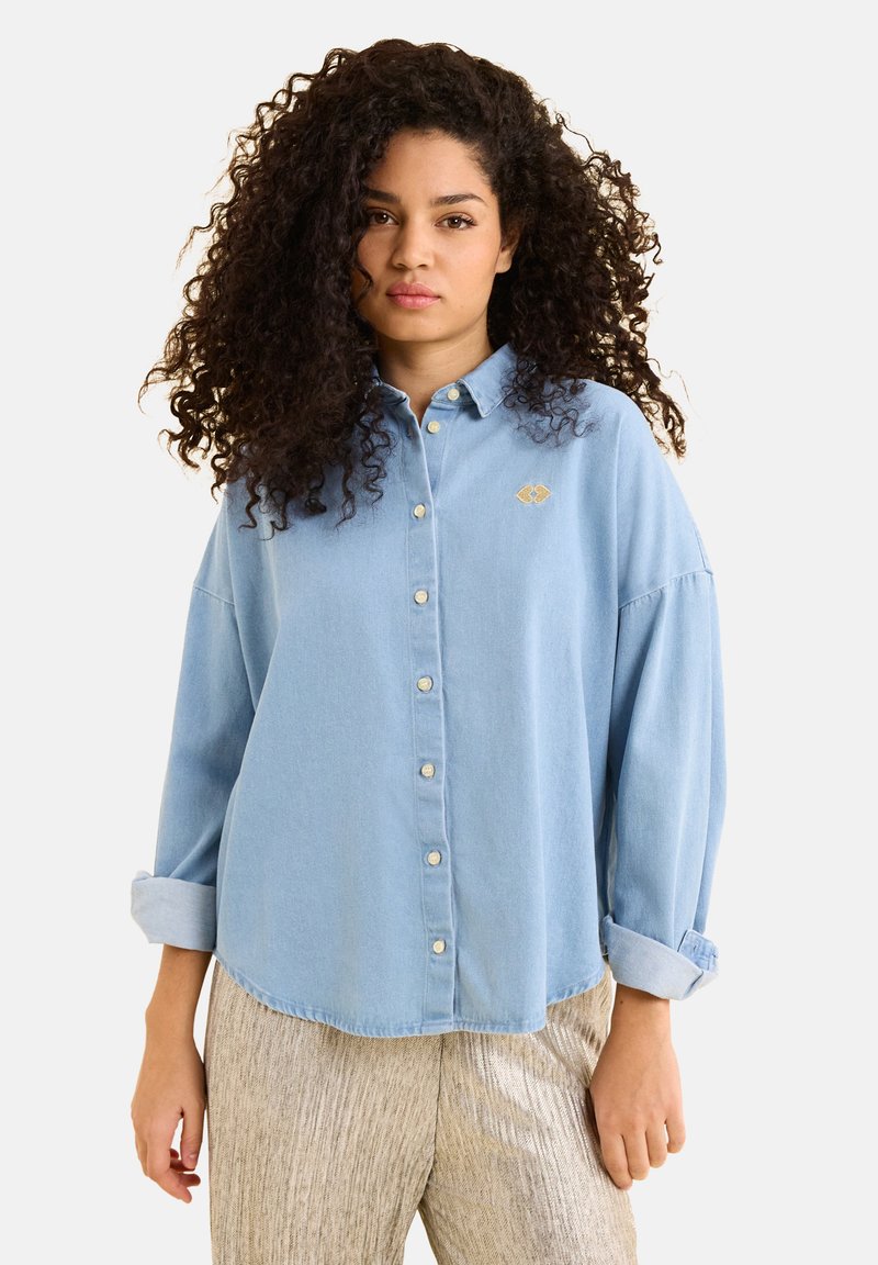 Camicia in denim azzurro chiaro con bottone, dal taglio morbido, maniche arrotolate e un piccolo dettaglio ricamato in oro sul petto.