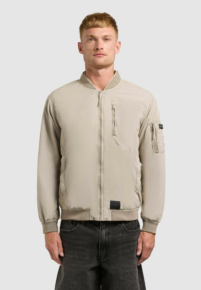 Jeune homme aux cheveux roux courts portant une veste bomber beige clair avec des poches zippées et un jean foncé, debout devant un fond uni.