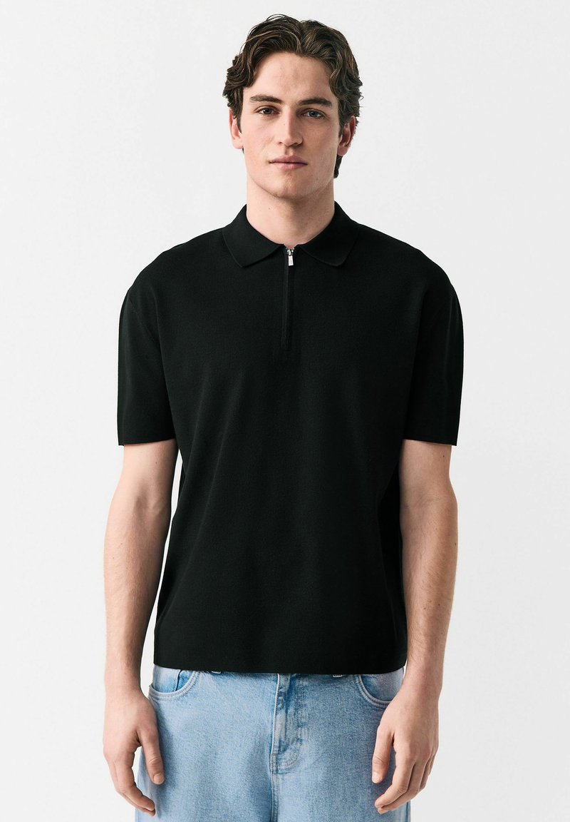 Sort polo t-shirt med korte ærmer og lynlås i halsen, lavet af tekstureret stof. Sættet er kombineret med lyseblå denim jeans.