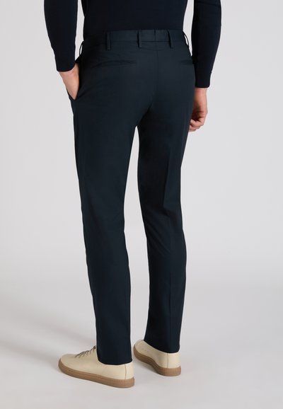 Uomo che indossa pantaloni slim fit blu navy e sneakers beige, in piedi con le mani in tasca contro uno sfondo chiaro.