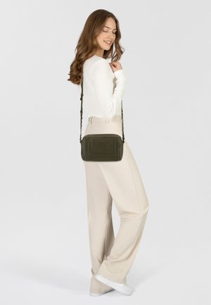 Olijfgroene leren schoudertas met gestikte details, rechthoekige vorm en een afneembare kettingriem. Model draagt een crème top en beige broek.