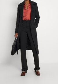 Zwarte lange jas over een rode satijnen blouse, gecombineerd met zwarte broek en bruine schoenen. Een gestructureerde zwarte tas wordt in één hand vastgehouden.