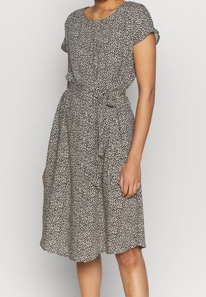 Femme portant une robe noire à manches courtes, longueur genou, avec un petit motif floral blanc et une ceinture nouée à la taille.