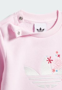 Světlounce růžová mikina s vyšitým stříbrným logem a barevnými květinovými detaily. Má zapínání na cvočky u límce a měkkou texturu látky.