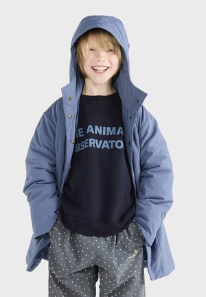 Blauwe capuchonjack met ritssluiting en drukknoopsluiting over een donkere sweatshirt met lichtblauwe tekst. Gecombineerd met grijze broek met stippen.