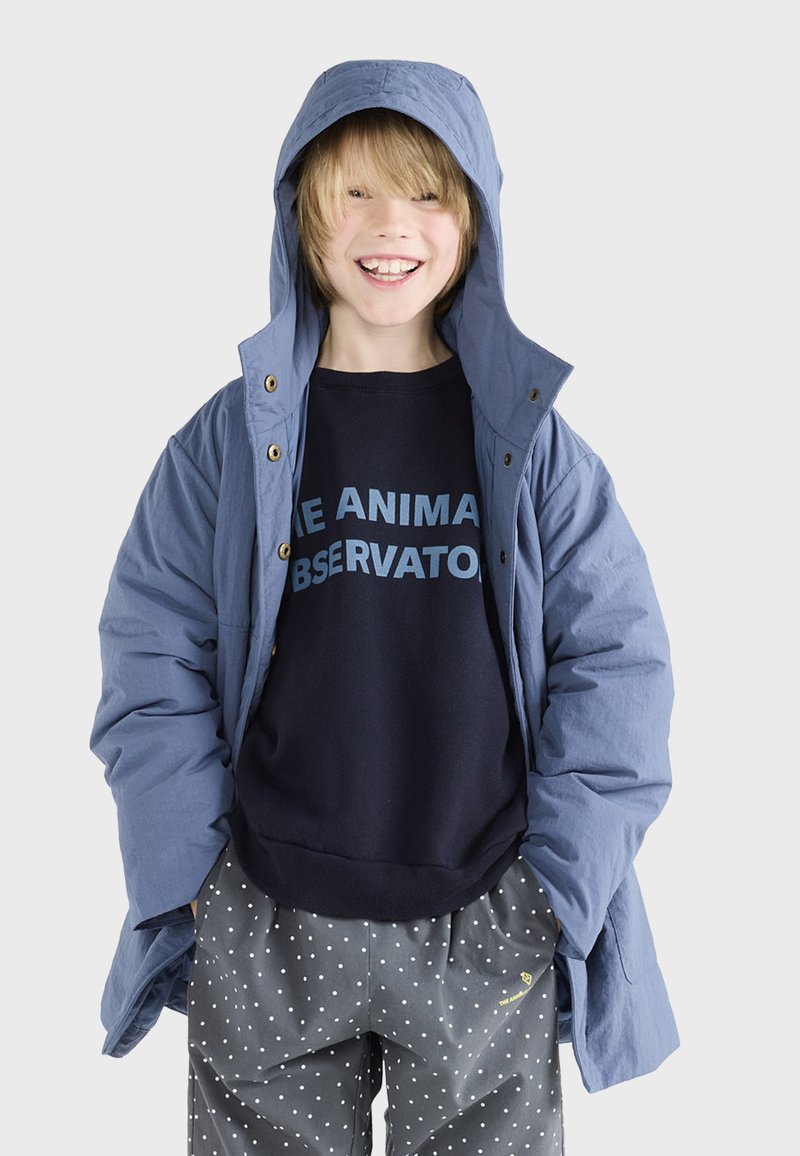 Blauer Kapuzenjacke mit Reißverschluss und Druckknopfverschluss über einem dunklen Sweatshirt mit hellblauer Schrift. Kombiniert mit grauen Polka-Dot-Hosen.