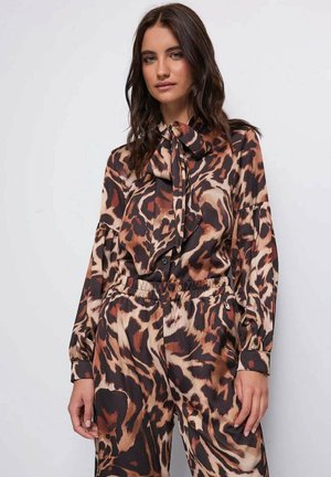 Donna con lunghi capelli castani che indossa una camicetta marrone e nera a stampa leopardata con fiocco e pantaloni abbinati con vita elastica.