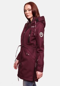 Burgundy waterproof jacket with a hood, drawstrings, multiple zippered pockets, logo on sleeve, and a smooth texture. Long, fitted design.