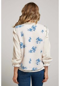 Femme aux cheveux ondulés portant un gilet en polaire blanc avec des taches florales bleues sur un chemisier crème et un jean bleu, vue de dos.