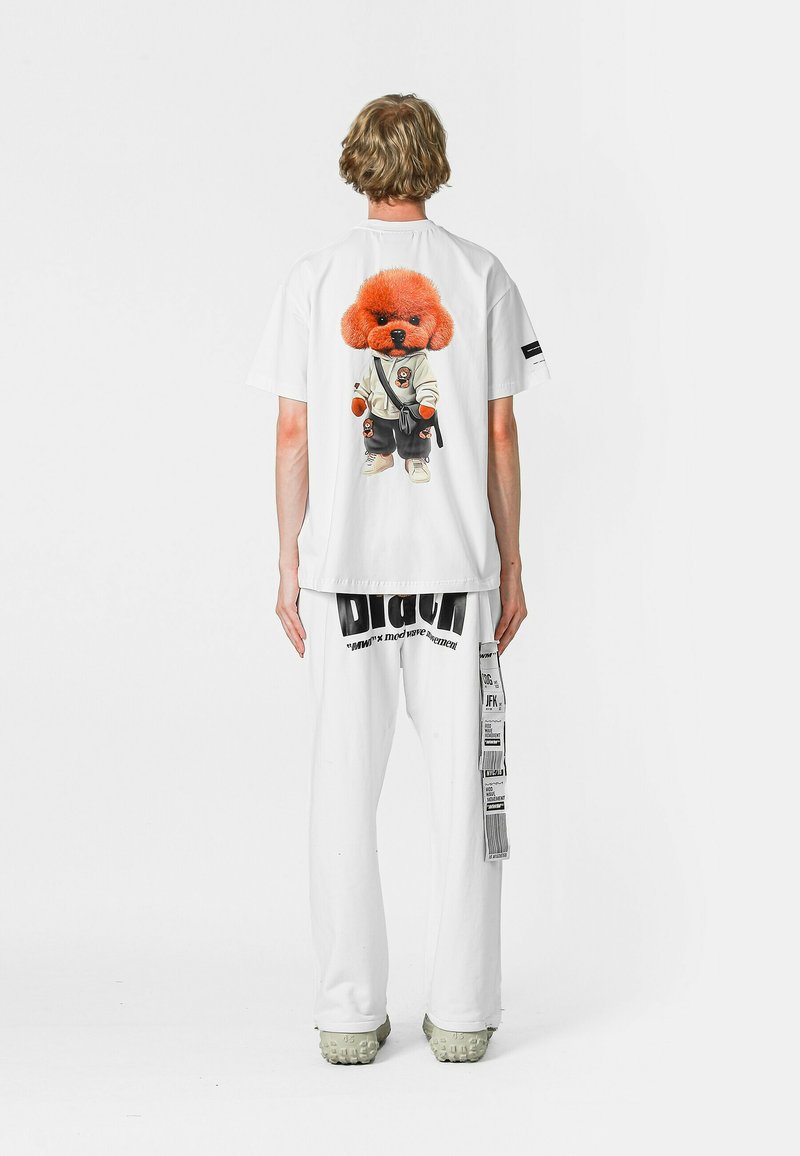 La camiseta blanca presenta un gráfico de un perro de dibujos animados con pelaje naranja, vistiendo un traje blanco, combinado con pantalones blancos que llevan texto y acentos en negro.