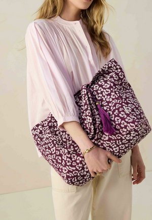 Purple patterned fabric tote bag with white abstract shapes, featuring a tassel detail. Model wearing a pleated light pink blouse.