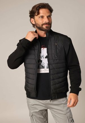 Veste noire avec un devant matelassé, des manches noires lisses et une poche poitrine zippée. Portée par-dessus un t-shirt graphique noir et un pantalon clair.