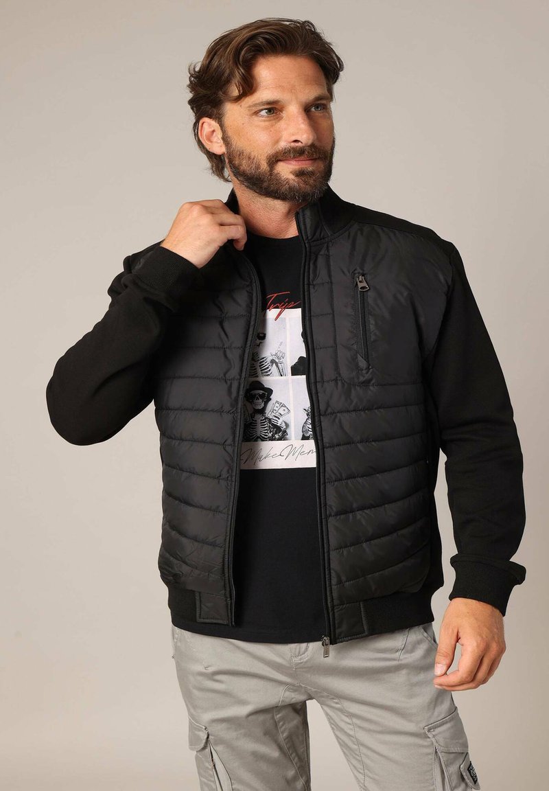 Zwarte jas met gewatteerde voorkant, gladde zwarte mouwen en een ritszak op de borst. Gemaakt boven een zwart T-shirt met grafisch ontwerp en lichte broek.