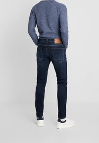 Jeans de color azul oscuro con corte ajustado, con una etiqueta de cuero en la parte trasera de la cintura y dos bolsillos traseros. Combinados con un suéter azul texturizado y zapatillas blancas.