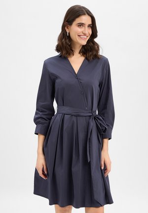 Kobieta uśmiechająca się, ubrana w ciemnoniebieską sukienkę z długim rękawem, z dekoltem w kształcie litery V, guzikami, plisowaną spódnicą i wiązanym paskiem w talii.