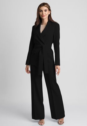 Kobieta stojąca w czarnym żakiecie z paskiem i szerokich spodniach z beżowymi butami na obcasie z odkrytymi palcami, na tle jednolitego jasnego tła.