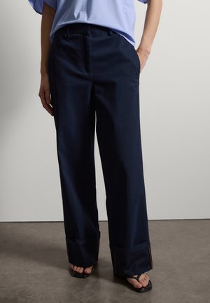 Person trägt marineblaue, weit geschnittene Hosen mit umgeschlagenen Säumen, ein hellblaues Oberteil und schwarze Sandalen mit offenen Zehen, steht auf einem grauen Boden.