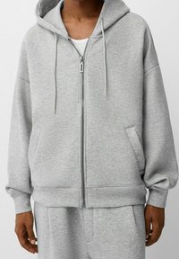 Sudadera con cremallera gris hecha de tela suave, con un ajuste relajado, capucha grande y dos bolsillos laterales. Incluye cordones y puños acanalados.