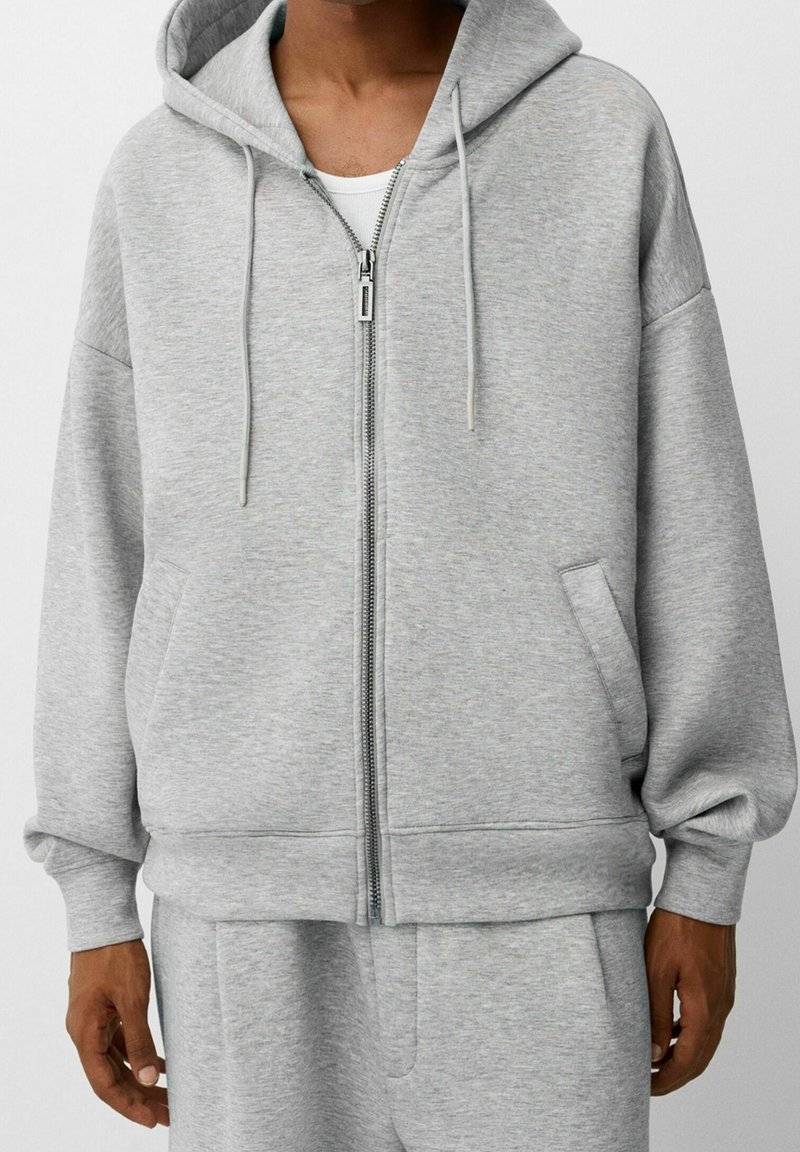 Sudadera con cremallera gris hecha de tela suave, con un ajuste relajado, capucha grande y dos bolsillos laterales. Incluye cordones y puños acanalados.