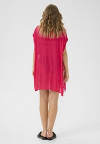 Túnica rosa transparente con mangas anchas, un corte holgado y aberturas laterales, combinada con sandalias negras de slip-on. La vista trasera muestra el cabello cayendo.