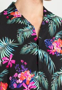 Maglietta nera a maniche corte con stampa floreale tropicale in colori vivaci, con foglie di palma verdi e vari fiori. Dettagli con bottoni visibili.