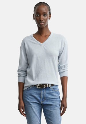 Femme aux cheveux tressés portant un pull bleu clair à col en V et un jean bleu avec une ceinture noire devant un fond uni.
