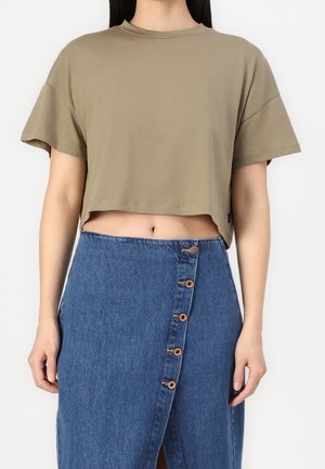 Kvinde iført en kort olivengrøn t-shirt og en blå denimnederdel med højtaljede knapper foran, stående foran en ensfarvet baggrund.