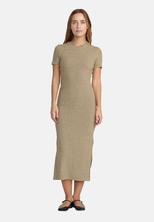 Femme debout portant une robe midi beige ajustée à manches courtes avec des chaussures Mary Jane noires sur un fond blanc uni.