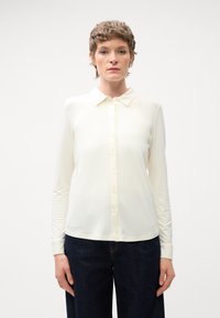 Bluza z dolgimi rokavi v svetli kremni barvi, z gumbom na sprednji strani in klasičnim ovratnikom, izdelana iz gladkega materiala z rahlo prilegnjeno silhueto.