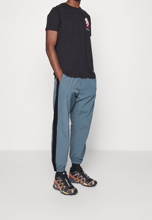 Man draagt een zwart T-shirt met een grafisch ontwerp, blauwe trainingsbroek met zwarte strepen aan de zijkant, en zwarte sportieve schoenen met oranje accenten, staand met handen in de zakken.