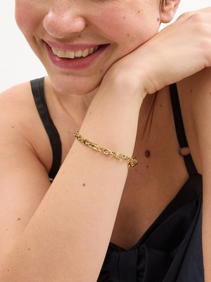 Uśmiechnięta kobieta w czarnym topie bez rękawów i złotej bransoletce łańcuchowej spoczywająca ramieniem blisko twarzy.