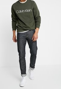 Felpa verde oliva con testo "Calvin Klein", indossata sopra una camicia bianca, abbinata a jeans in denim scuro e sneakers bianche.