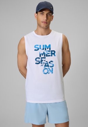 Mężczyzna w granatowej czapce, białej koszulce bez rękawów z napisem „SUMMER SEASON” i jasnoniebieskich szortach na jednolitym szarym tle.