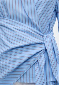 Chemise bleue avec fines rayures verticales blanches et marron nouée à la taille.