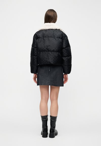 Γυναίκα που στέκεται με την πλάτη στραμμένη, φοράει μαύρο μπουφάν puffer με λευκή γούνινη γιακά, σκούρα μίνι φούστα και μαύρες δερμάτινες μπότες μέχρι τον μέσο της γάμπας.