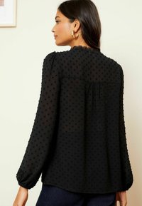 Blusa nera con motivo a pois testurizzati, maniche lunghe, cuciture delle spalle arricciate e bordo in pizzo al colletto. Materiale liscio e leggero.
