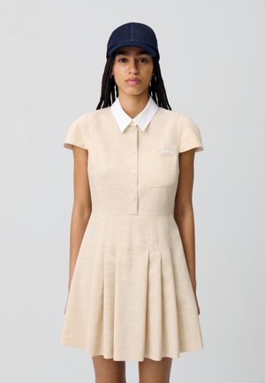 Abito plissettato beige con maniche corte, colletto bianco e chiusura con bottoni. Include una tasca piccola con un'etichetta. Indossato con un berretto scuro.