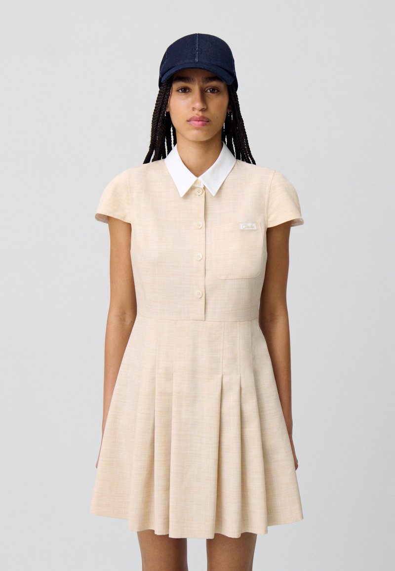 Robe plissée beige à manches courtes, avec un col blanc et une fermeture à boutons. Comprend une petite poche avec une étiquette. Portée avec une casquette sombre.