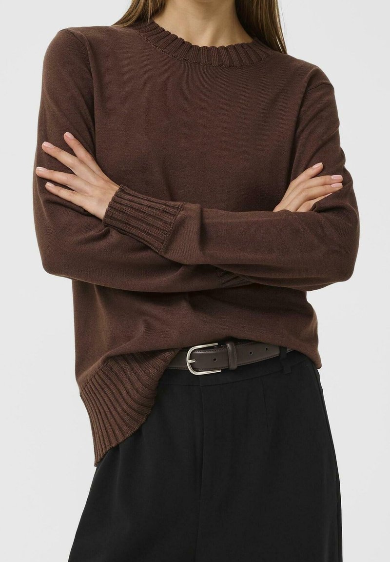 Femme aux bras croisés portant un pull marron côtelé rentré dans un pantalon noir avec une ceinture foncée, sur un fond clair uni.