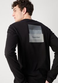 Melnā garā piedurkņu T-krekls ar caurspīdīgu taisnstūra virsējo slāni, uz kura augšējā daļā ir uzraksts "Calvin Klein". Gluda kokvilnas audums.