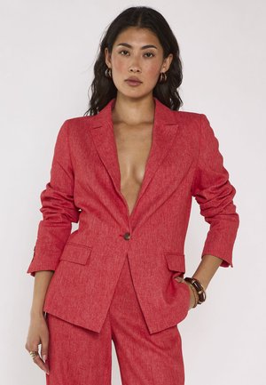Vrouw met lang donker haar draagt een rode textuurblazer en bijpassende broek, met één hand in de zak en een neutrale gezichtsuitdrukking.