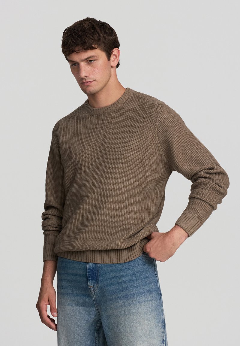 Maglione brown a maglia texturizzata con scollatura a girocollo e polsini a coste, abbinato a jeans chiari e sbiaditi. Il tessuto appare caldo e informale.