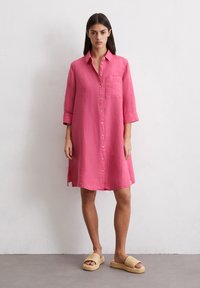 Kreklu kleita spilgti rozā krāsā, ar garām piedurknēm, krūšu kabatu un sānu šķēlumiem. Stilizēta ar adītām bēšām sandālēm.