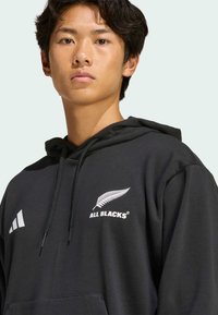 Schwarze Kapuzenjacke mit Kängurutasche, ausgestattet mit dem weißen "ALL BLACKS"-Logo und einem Farn-Design. Hergestellt aus weichem Material mit verstellbarem Kordelzug an der Kapuze.