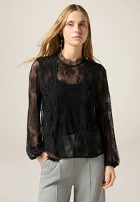 Zwarte kanten blouse met lange mouwen, bloemenpatronen en een geschulpte kraag. Daaronder is een zwarte tanktop zichtbaar; gecombineerd met grijze broek.