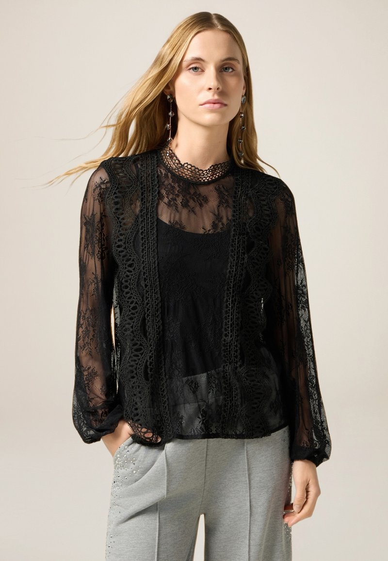 Zwarte kanten blouse met lange mouwen, bloemenpatronen en een geschulpte kraag. Daaronder is een zwarte tanktop zichtbaar; gecombineerd met grijze broek.